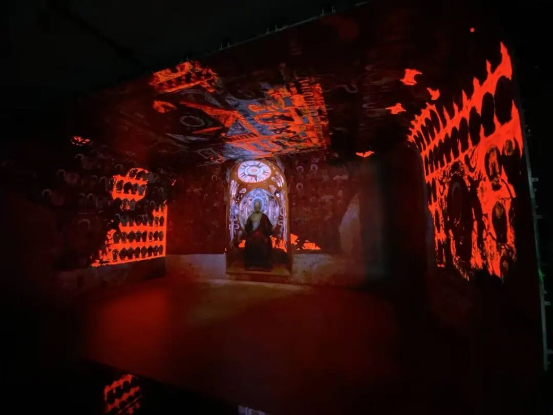 Inside the immersive projection cave, warm red tones recreate Dunhuang cave murals, thousand Buddha figures, and a central niche. Inside the immersive projection cave, warm red tones recreate Dunhuang cave murals, thousand Buddha figures, and a central niche.